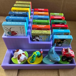 May include: A purple tiered display case holding miniature children's books and toy figures. The books have colorful covers with titles like "The Very Busy Spider" and "The Very Hungry Caterpillar." Toy figures include a puppy, shark, and a red truck.