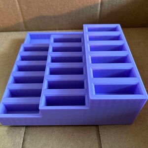 May include: A lavender-colored, tiered organizer with multiple rectangular compartments. The organizer has a stepped design, with three distinct levels of storage. The compartments are uniform in size and shape, suitable for small items.