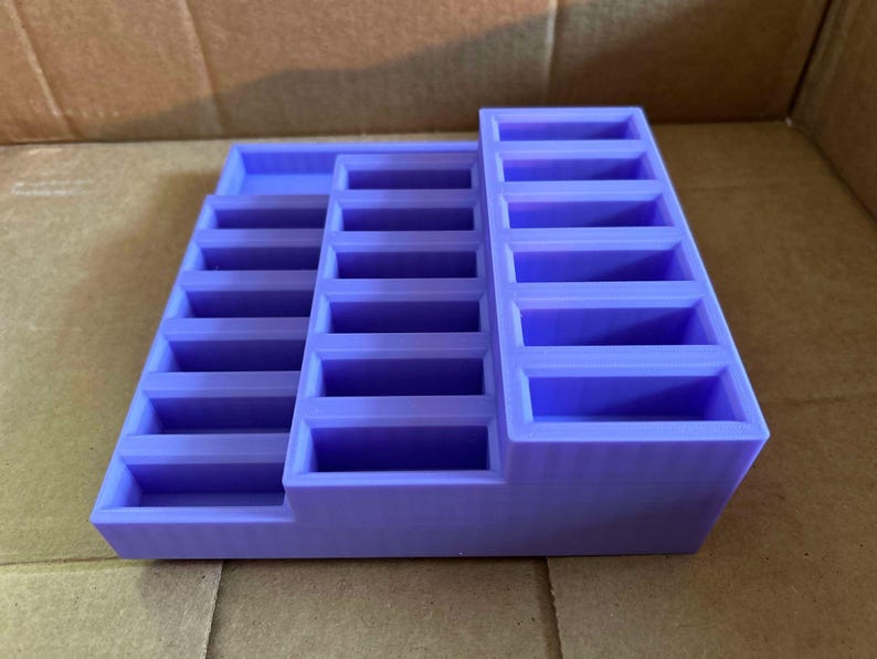 May include: A lavender-colored, tiered organizer with multiple rectangular compartments. The organizer has a stepped design, with three distinct levels of storage. The compartments are uniform in size and shape, suitable for small items.