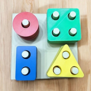 May include: A wooden toy with five different shapes: a circle, a square, a rectangle, a triangle, and a diamond. Each shape has a hole in the center and a white peg that can be inserted into the hole.