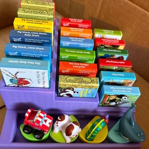May include: A purple display case filled with colorful children's books and small toy figures. The books feature titles like "Big Shark, Little Shark" and "The Very Hungry Caterpillar." Toy figures include a red truck, a puppy, and a shark.