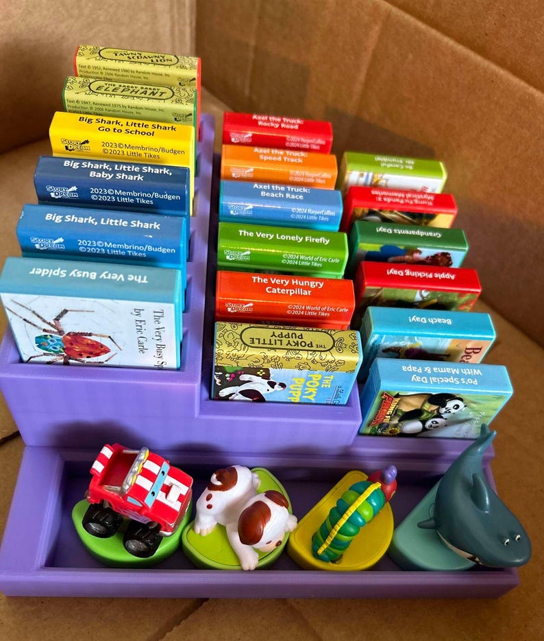May include: A purple display case filled with colorful children's books and small toy figures. The books feature titles like "Big Shark, Little Shark" and "The Very Hungry Caterpillar." Toy figures include a red truck, a puppy, and a shark.