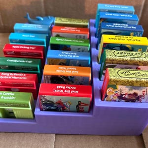 May include: A purple tiered organizer filled with colorful children's books. Titles include "Axel the Truck: Rocky Road", "Tawny Scrawny Lion", and "Big Shark, Little Shark, Go to School". A blue whale figurine sits on top.