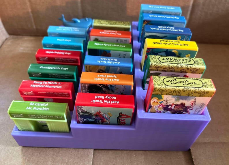 May include: A purple tiered organizer filled with colorful children's books. Titles include "Axel the Truck: Rocky Road", "Tawny Scrawny Lion", and "Big Shark, Little Shark, Go to School". A blue whale figurine sits on top.