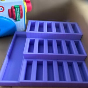 May include: A purple plastic organizer with three tiered sections and a separate tray. A blue and red Little Tikes toy camera is in the background. The organizer is designed to hold small items.