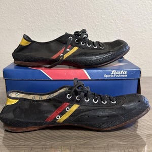 Könnte beinhalten: Paar schwarze Vintage-Bata-Leinenschuhe mit roten und gelben Streifen. Die Sneaker haben weiße Schnürsenkel und eine braune Sohle. Sie stehen auf einer blauen Bata Sports Footwear-Box.