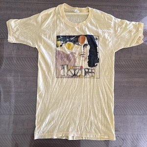 May include: A pale yellow t-shirt with a rectangular graphic featuring a portrait and the band name "The Doors". The graphic includes a face with long black hair and an orange circle. The shirt has short sleeves and a crew neck.