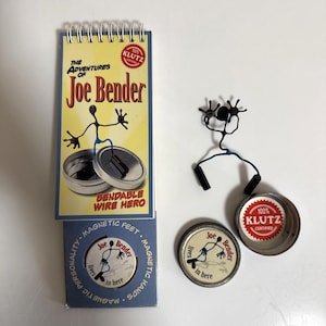 May include: A novelty toy set featuring a bendable wire figure named Joe Bender. The set includes a notepad, magnetic feet, and hands, along with two metal containers. The packaging displays the text "The Adventures of Joe Bender."