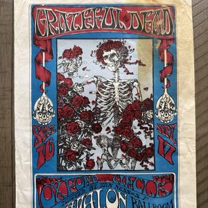 May include: Vintage concert poster for the Grateful Dead, featuring a skeleton adorned with roses. The poster has a blue and red color scheme with the band's name at the top. The text includes the venue, date, and ticket outlets.