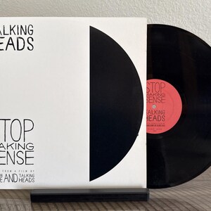 May include: A Talking Heads Stop Making Sense vinyl record album cover with a white background, black text, and a large black semicircle. A black vinyl record with a pink center label is partially pulled out of the white cardboard sleeve.