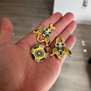 K&ouml;nnte beinhalten: Drei Emaille-Anstecknadeln mit goldfarbenen Metallr&auml;ndern. Zwei Anstecknadeln sind wie K&auml;fer mit schwarzen, gelben und wei&szlig;en Fl&uuml;geln geformt, eine zeigt einen roten Kreis mit wei&szlig;en Punkten, die andere eine blaue Form. Die dritte ist kreisf&ouml;rmig.