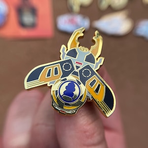 K&ouml;nnte beinhalten: Eine goldfarbene Emaille-Anstecknadel in Form eines stilisierten K&auml;fers mit gro&szlig;en Mandibeln. Das Design zeigt schwarze, orangefarbene und wei&szlig;e Bereiche mit einem kreisf&ouml;rmigen blau-wei&szlig;en Emblem in der K&ouml;rpermitte, akzentuiert durch goldene Konturen.