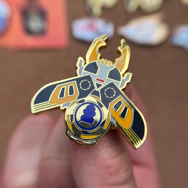 K&ouml;nnte beinhalten: Eine goldfarbene Emaille-Anstecknadel in Form eines stilisierten K&auml;fers mit gro&szlig;en Mandibeln. Das Design zeigt schwarze, orangefarbene und wei&szlig;e Bereiche mit einem kreisf&ouml;rmigen blau-wei&szlig;en Emblem in der K&ouml;rpermitte, akzentuiert durch goldene Konturen.