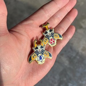 K&ouml;nnte beinhalten: Zwei Metall-Emaille-Anstecknadeln in Form von Nashornk&auml;fern mit goldfarbenen Umrandungen. Die Designs zeigen schwarze, wei&szlig;e und gelbe Fl&uuml;gel mit geometrischen Mustern. Eine Anstecknadel zeigt ein rotes Kreissymbol, die andere eine blaue Silhouette.