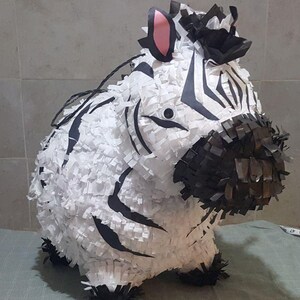 Puede incluir: Una piñata con forma de cebra blanca y negra con una melena negra y una oreja rosa. La piñata está hecha de papel de seda y está lista para llenarse de golosinas.