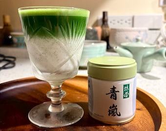 May include: A crystal glass filled with a layered green matcha drink featuring a vibrant green top and a creamy white base, positioned next to a cylindrical metal tin with a light green lid and Japanese text reading Aoarashi.