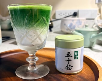 May include: A faceted glass filled with a vibrant green matcha latte layered over white milk, positioned next to a cylindrical metal tin with a light green lid. The tin features white labels with black Japanese characters and the text ISUZU.