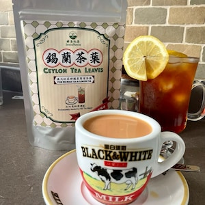 May include: A close-up of a white ceramic mug with a black and white cow design and the text "Black & White" and "Milk". The mug is filled with a light brown liquid and is sitting on a white saucer. A bag of tea leaves is in the background.