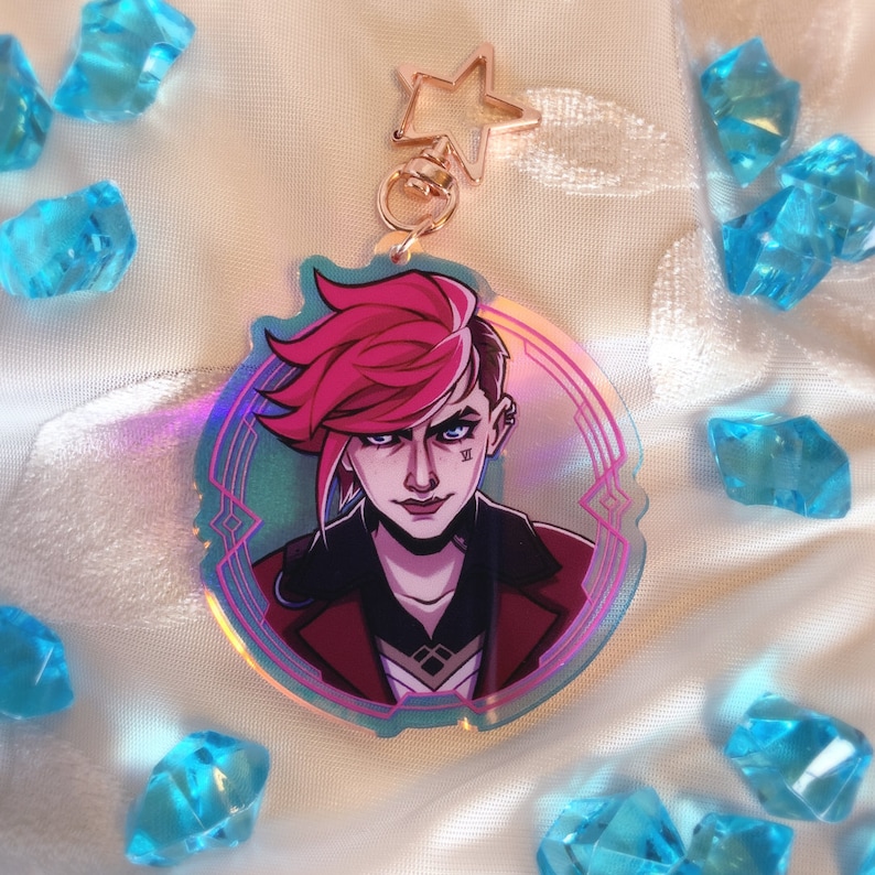 May include: A holographic acrylic keychain featuring a character with pink hair and a stern expression. The keychain has a star-shaped clasp and a circular design with a teal and purple border. The character is wearing a red jacket.