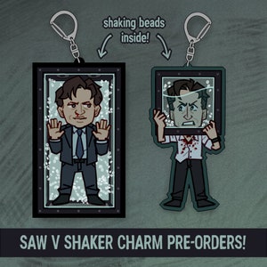 May include: Two Saw V shaker charms with a rectangular design and a silver keychain. One charm features a man in a suit, the other a man in a white shirt with blood. The text says "shaking beads inside!" and "SAW V SHAKER CHARM PRE-ORDERS!"