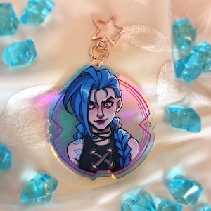 May include: A holographic acrylic keychain featuring a cartoon character with blue hair and pink eyes. The character is wearing a black top and has a tattoo on their arm. The keychain has a star-shaped clasp.