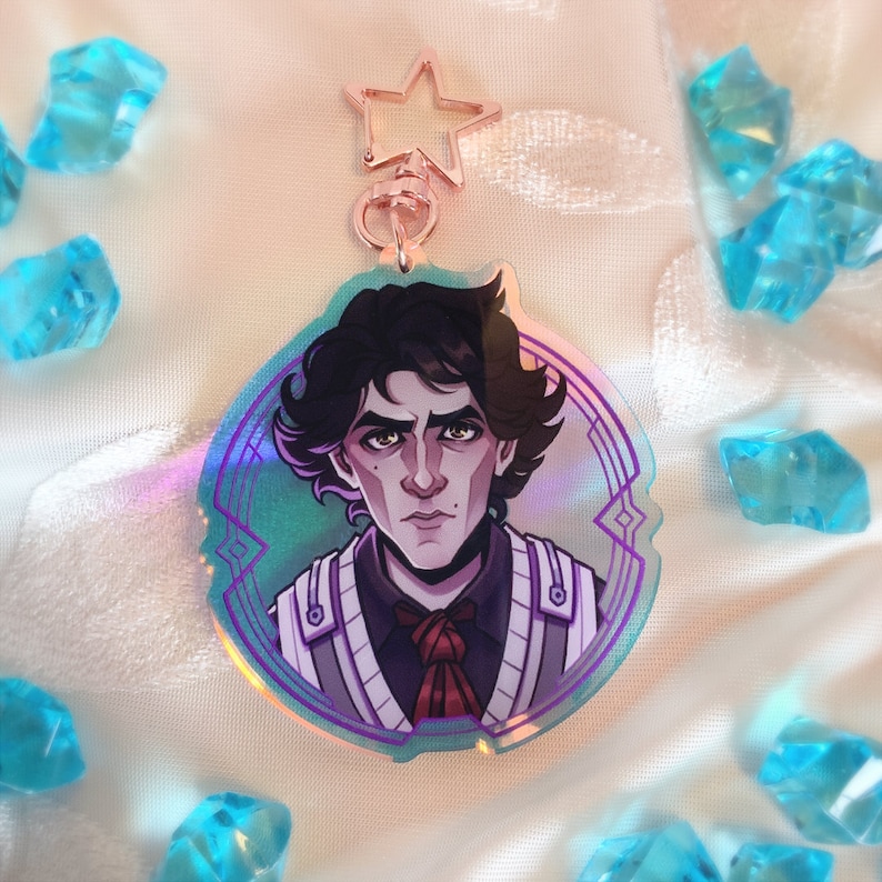 May include: A holographic acrylic charm featuring a stylized illustration of a person with dark hair and a stern expression. The charm has a rose gold star-shaped clasp and is surrounded by blue crystal-like objects.