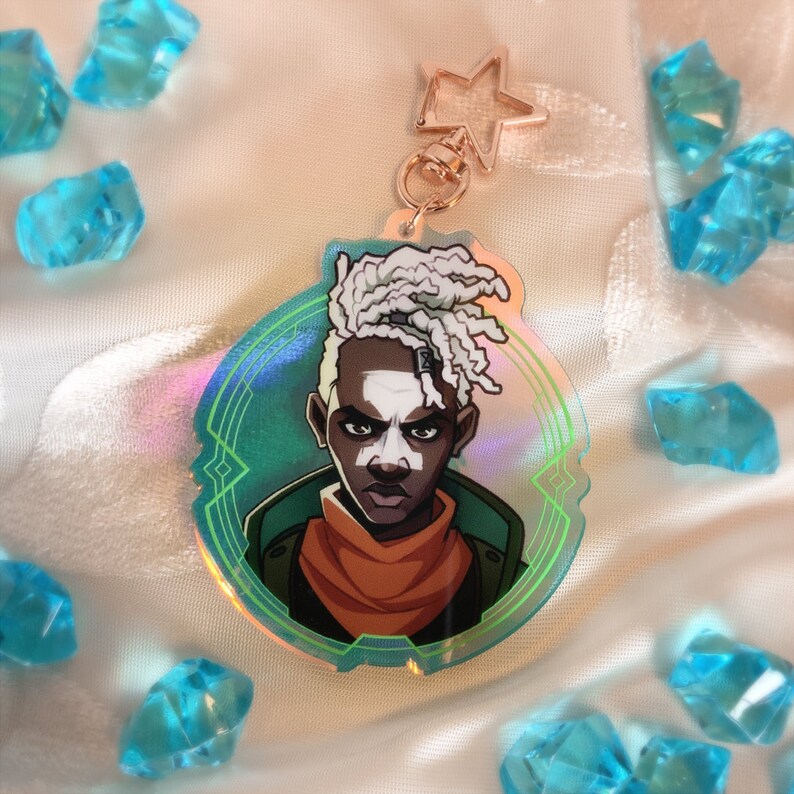 May include: A holographic keychain featuring a stylized illustration of a person with white dreadlocks, an orange scarf, and a green jacket. The keychain has a star-shaped clasp and is surrounded by blue crystal-like objects.