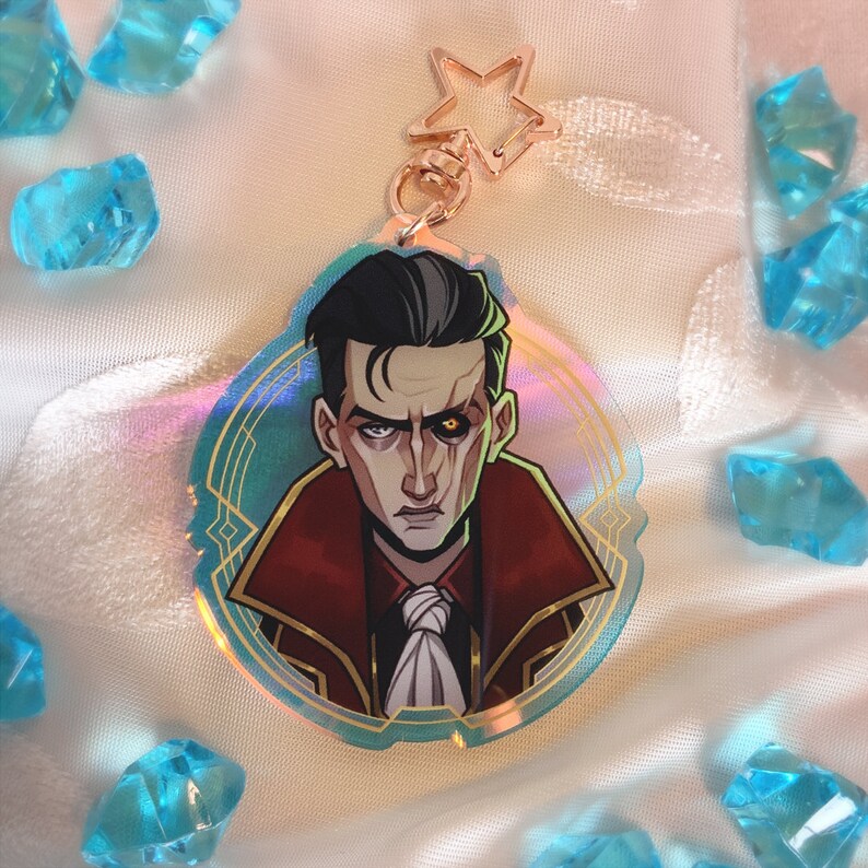 May include: A clear acrylic keychain featuring a stylized illustration of a character with dark hair, a red coat, and a stern expression. The keychain has a gold-colored star-shaped clasp and a holographic effect. The background includes blue crystal-like objects.