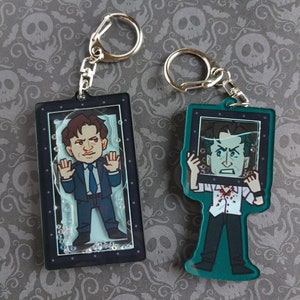 May include: Two keychains featuring cartoon illustrations of a man in a suit and another in a white shirt with blood. The keychains have silver metal rings and are encased in rectangular frames with a clear background and decorative bubbles.