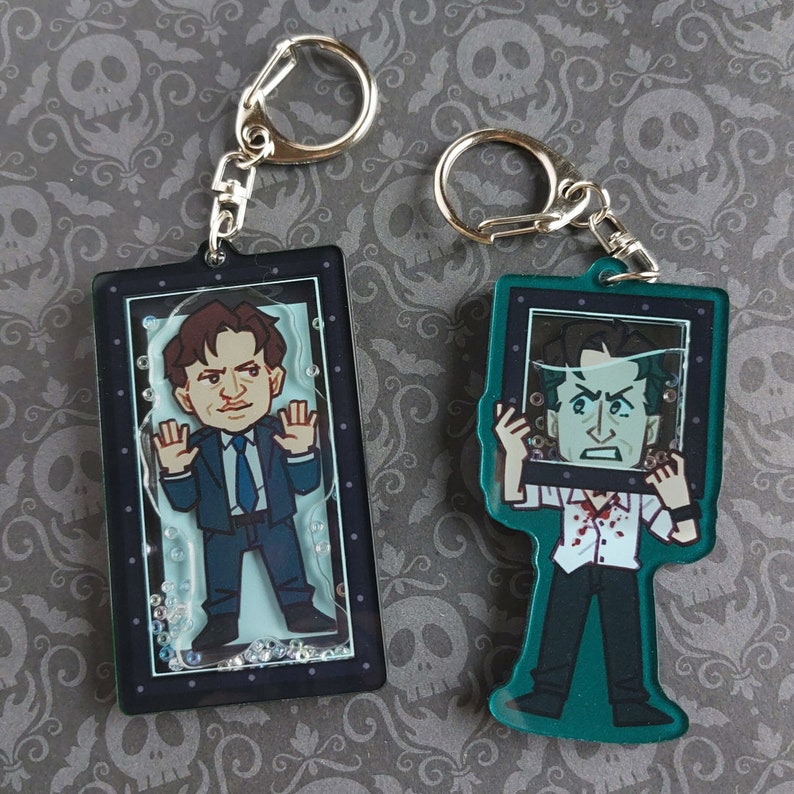 May include: Two keychains featuring cartoon illustrations of a man in a suit and another in a white shirt with blood. The keychains have silver metal rings and are encased in rectangular frames with a clear background and decorative bubbles.