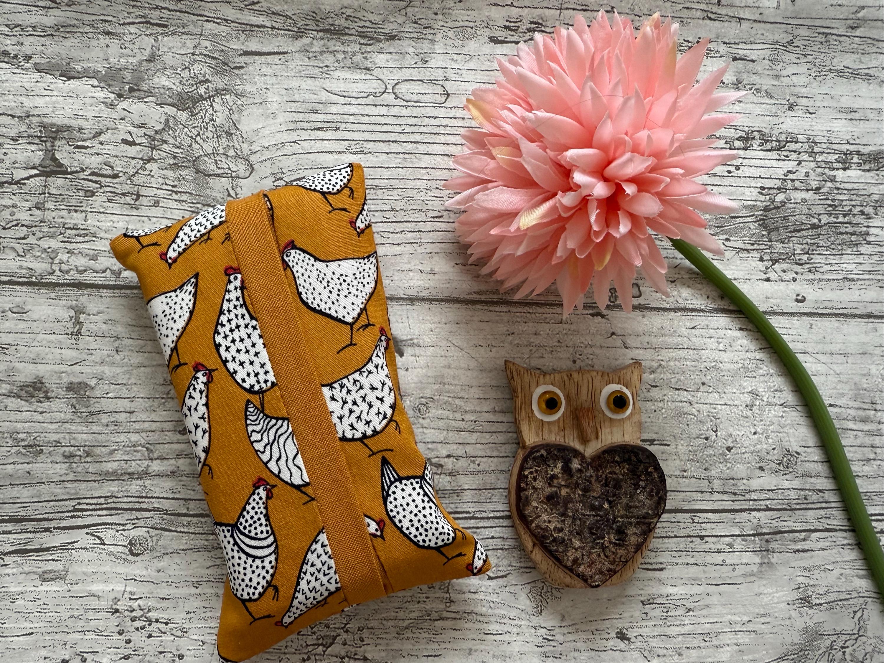 May include: Mustard yellow tissue box cover with a white chicken pattern. A wooden owl figurine with a heart-shaped centre and a pink flower are also in the image. The items are on a wooden surface.