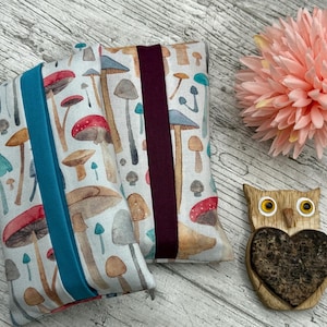 May include: Two rectangular fabric cases with a mushroom design, one with a teal band and the other with a claret band. A pink flower and a wooden owl figurine with a heart-shaped cutout are also present.