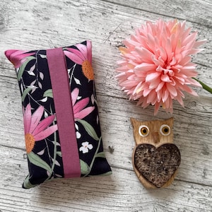 May include: A rectangular fabric pouch featuring a black background with a pink and green floral pattern and a solid mauve vertical band, accompanied by a light pink artificial flower and a small wooden owl ornament with a dark heart-shaped belly.
