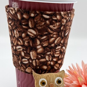May include: A burgundy coffee cup with a coffee bean print sleeve. The cup has a white lid. A wooden owl ornament and a pink flower are in the foreground.