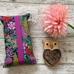 May include: A floral print tissue box cover with a magenta band, a pink flower, and a wooden owl figurine with a heart-shaped chest. The tissue box cover has a dark blue background with colorful flowers.