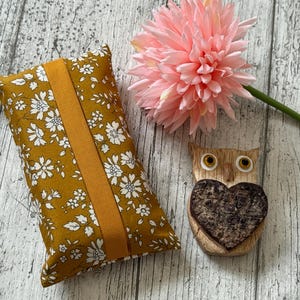 May include: Mustard-yellow tissue box cover with a white floral pattern and a matching centre band. A pink flower and a wooden owl figurine with a heart-shaped chest are also in the image.