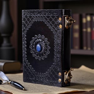 May include: Black leather journal with ornate embossed floral designs and a central blue gemstone. The journal features a clasp closure and is displayed with a pen and paper, suggesting a writing theme.