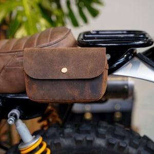 May include: A brown leather motorcycle saddlebag with a flap and gold-colored button closure. The bag is mounted on a brown leather motorcycle seat. The motorcycle has a yellow and black frame, and a black tire.