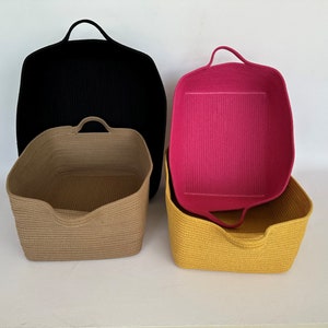 K&ouml;nnte beinhalten: Vier gewebte Aufbewahrungsk&ouml;rbe in Schwarz, Beige, Gelb und Pink. Die K&ouml;rbe haben eine rechteckige Form mit abgerundeten Ecken und integrierten Griffen. Der schwarze Korb ist der gr&ouml;&szlig;te, mit dem rosa Korb im gelben Korb.