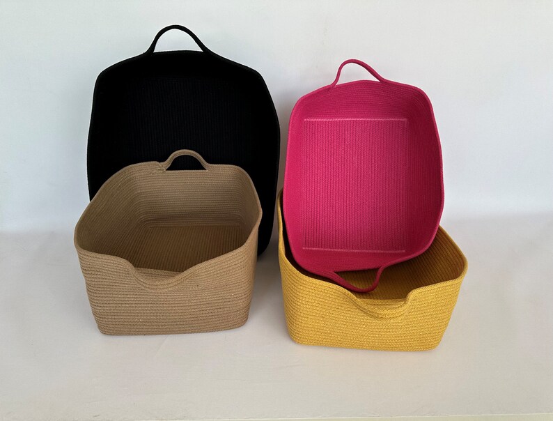 K&ouml;nnte beinhalten: Vier gewebte Aufbewahrungsk&ouml;rbe in Schwarz, Beige, Gelb und Pink. Die K&ouml;rbe haben eine rechteckige Form mit abgerundeten Ecken und integrierten Griffen. Der schwarze Korb ist der gr&ouml;&szlig;te, mit dem rosa Korb im gelben Korb.
