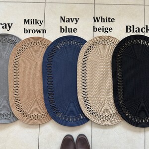 May include: Five oval-shaped braided rugs in different colors: gray, brown, navy blue, beige, and black. The rugs have a decorative braided border.