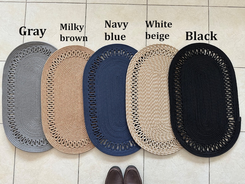 May include: Five oval-shaped braided rugs in different colors: gray, brown, navy blue, beige, and black. The rugs have a decorative braided border.