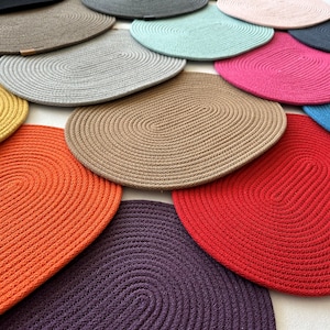 Könnte beinhalten: Eine Sammlung ovaler, gewebter Tischsets in verschiedenen Farben, darunter Rot, Orange, Gelb, Braun, Grau und Blau. Die Tischsets sind so angeordnet, dass ihre Textur und ihr Design zur Geltung kommen. Jedes Tischset ist etwa 30 cm lang.