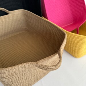 K&ouml;nnte beinhalten: Eine Sammlung rechteckiger Aufbewahrungsk&ouml;rbe in verschiedenen Farben. Der vordere Korb ist beige mit Griffen, die anderen sind schwarz, pink und gelb. Die K&ouml;rbe haben eine gewebte Textur.