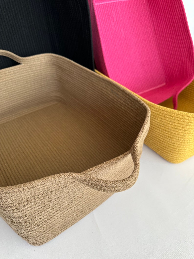 K&ouml;nnte beinhalten: Eine Sammlung rechteckiger Aufbewahrungsk&ouml;rbe in verschiedenen Farben. Der vordere Korb ist beige mit Griffen, die anderen sind schwarz, pink und gelb. Die K&ouml;rbe haben eine gewebte Textur.