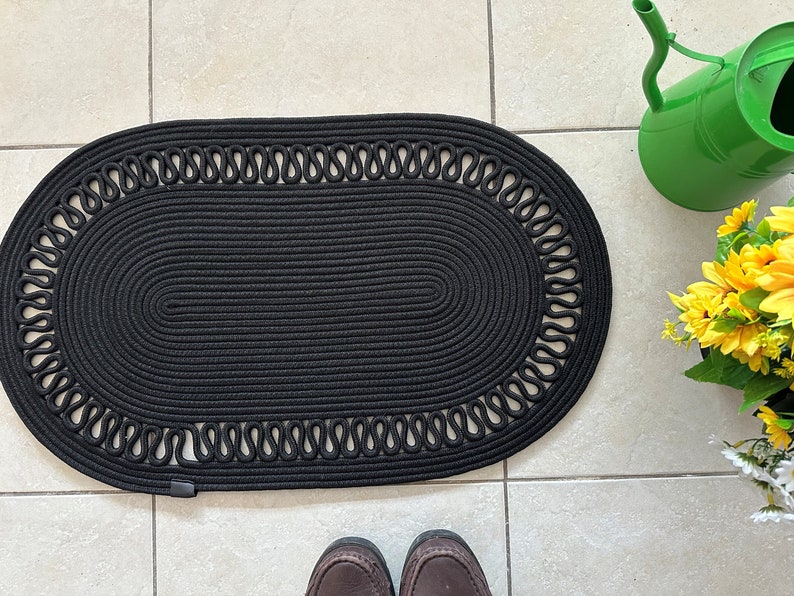 May include: Black oval braided rug with a decorative rope border. The rug is made of thick, woven rope and has a subtle texture.