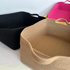 K&ouml;nnte beinhalten: Vier rechteckige Aufbewahrungsk&ouml;rbe in Schwarz, Beige und Rosa. Jeder Korb hat ein geflochtenes Seildesign und integrierte Griffe. Die K&ouml;rbe sind leer und scheinen aus einem weichen, flexiblen Material zu bestehen.