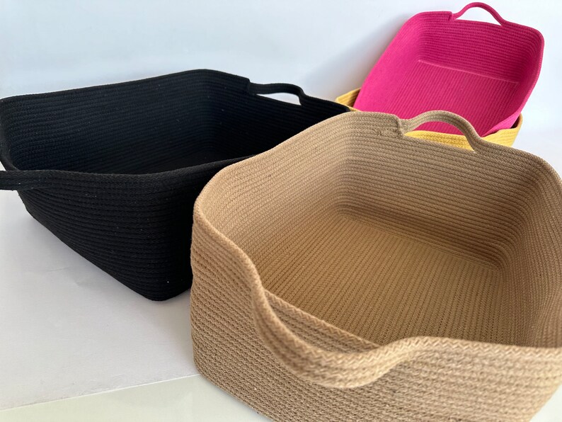 K&ouml;nnte beinhalten: Vier rechteckige Aufbewahrungsk&ouml;rbe in Schwarz, Beige und Rosa. Jeder Korb hat ein geflochtenes Seildesign und integrierte Griffe. Die K&ouml;rbe sind leer und scheinen aus einem weichen, flexiblen Material zu bestehen.