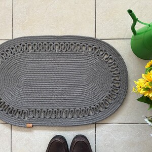 May include: A gray oval braided rug with a decorative border. The rug is on a tiled floor.