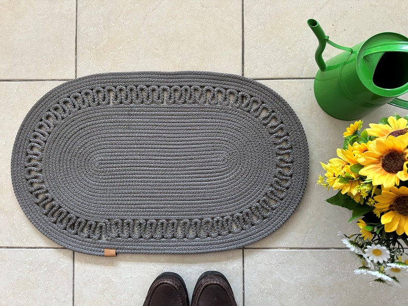 May include: A gray oval braided rug with a decorative border. The rug is on a tiled floor.